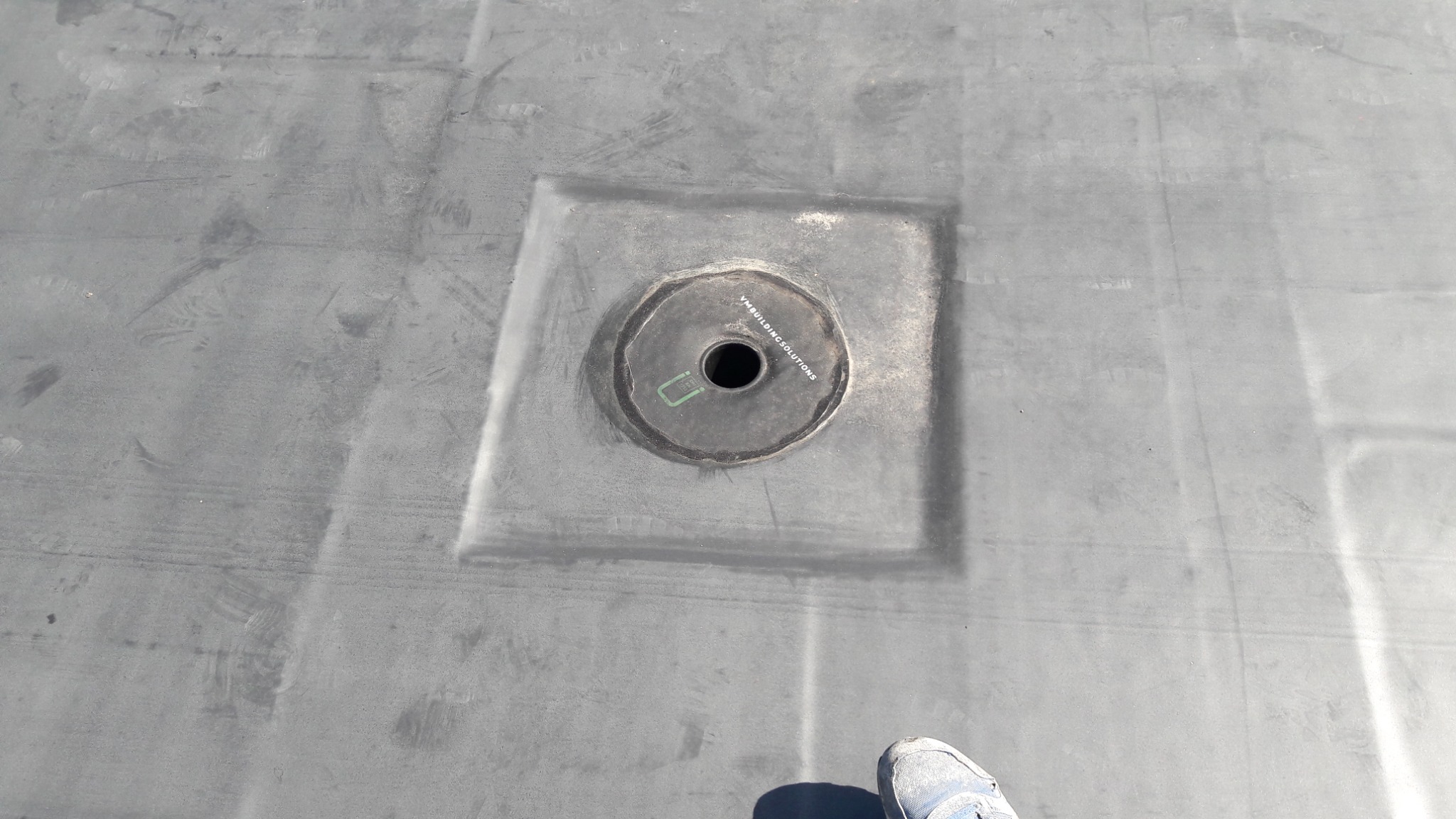 Szare pokrycie dachu z odpływem wody deszczowej, widoczne ślady montażu i fragment buta w prawym dolnym rogu.