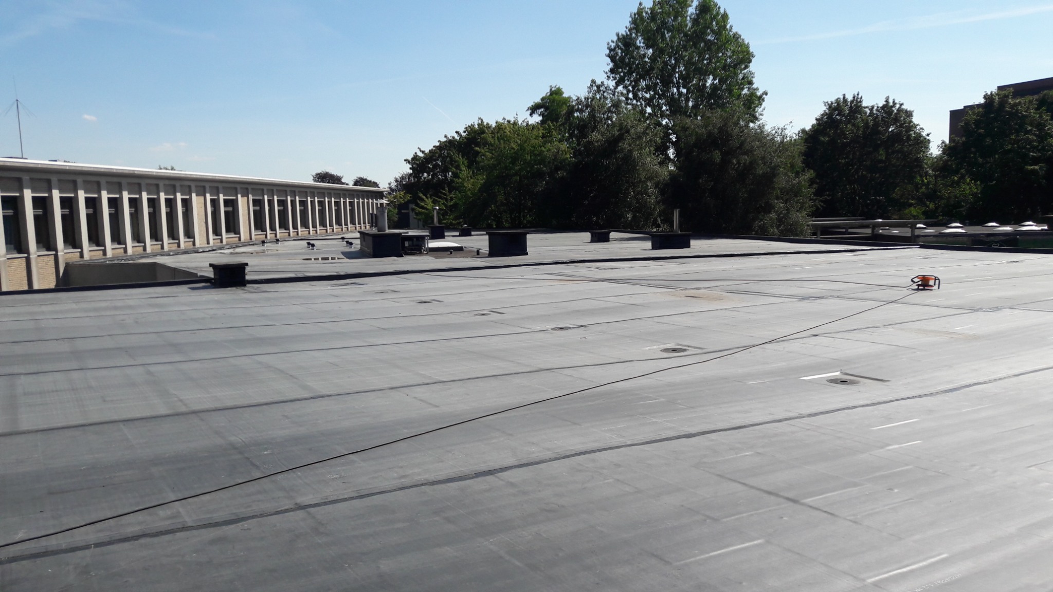 Płaski dach budynku usługowego pokryty szarą membraną bitumiczną, widoczne kominki wentylacyjne i przewód elektryczny podłączony do pomarańczowego urządzenia. Budynek z rzędem okien w tle.