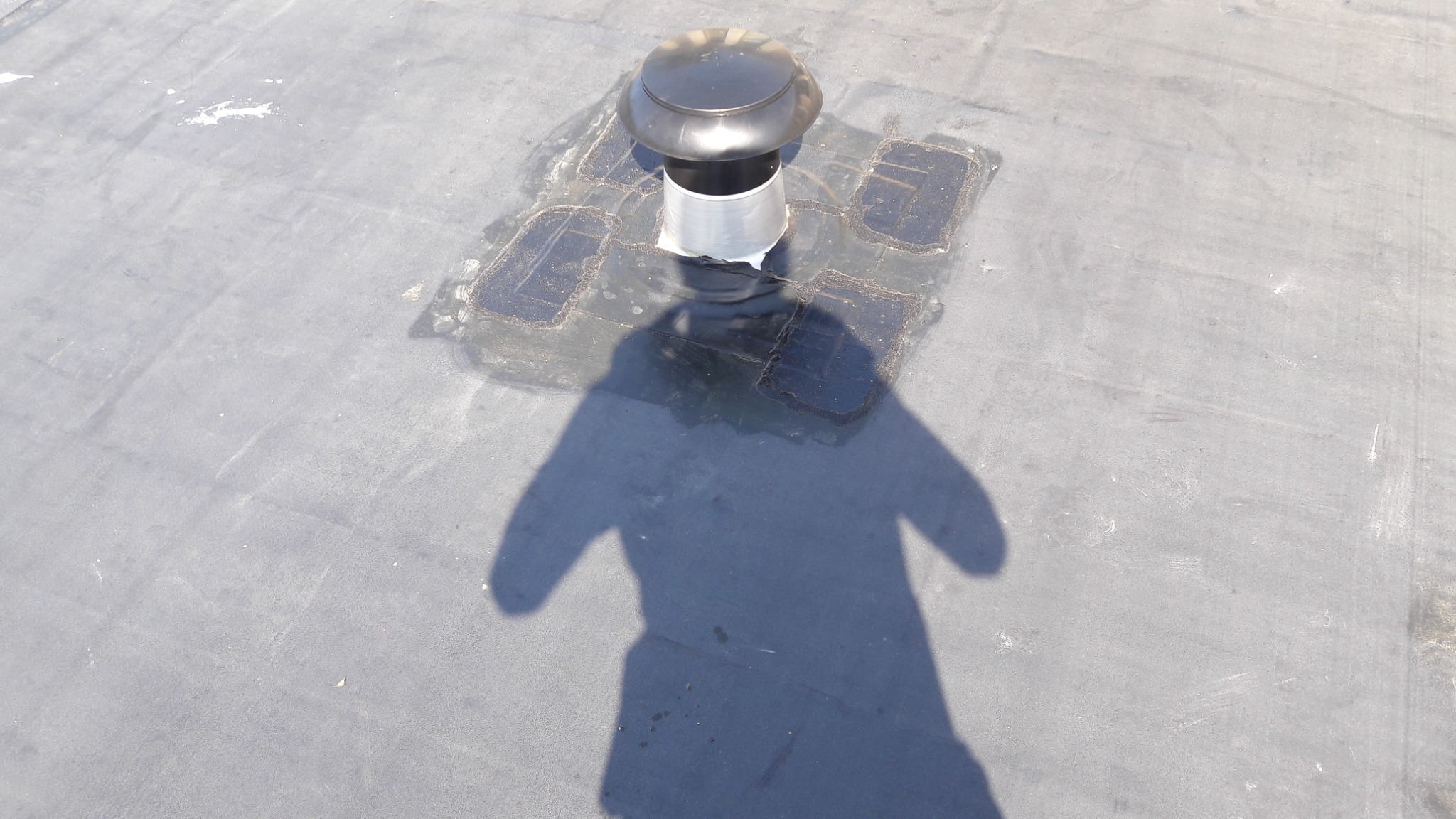 Wentylacja kominowa na płaskim dachu pokrytym papą, z widocznymi śladami napraw i uszczelnien wokół podstawy kominka wentylacyjnego, cień osoby fotografującej.