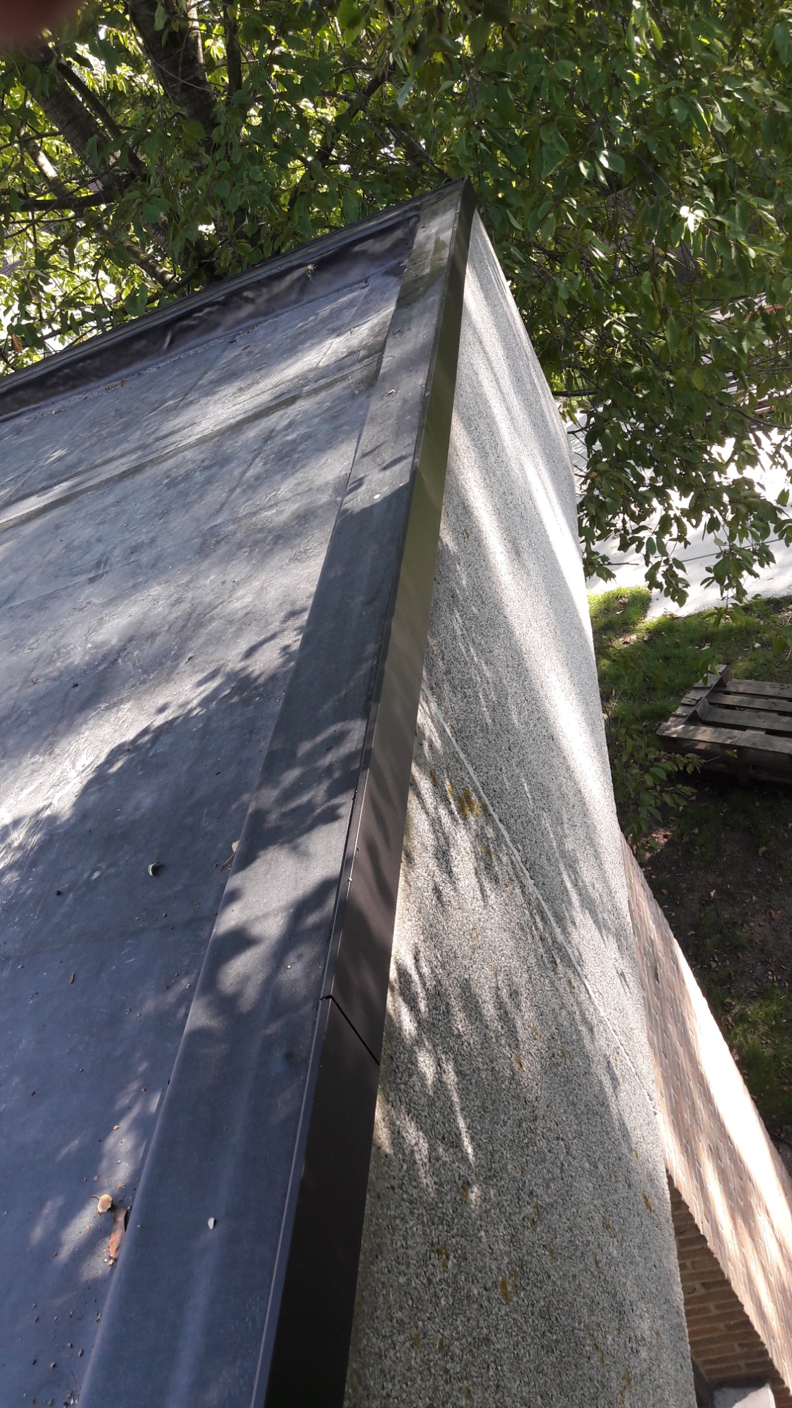 Płaski dach pokryty papą z obróbką blacharską, widok z góry na krawędź dachu i ścianę budynku z widocznymi cieniami drzew.