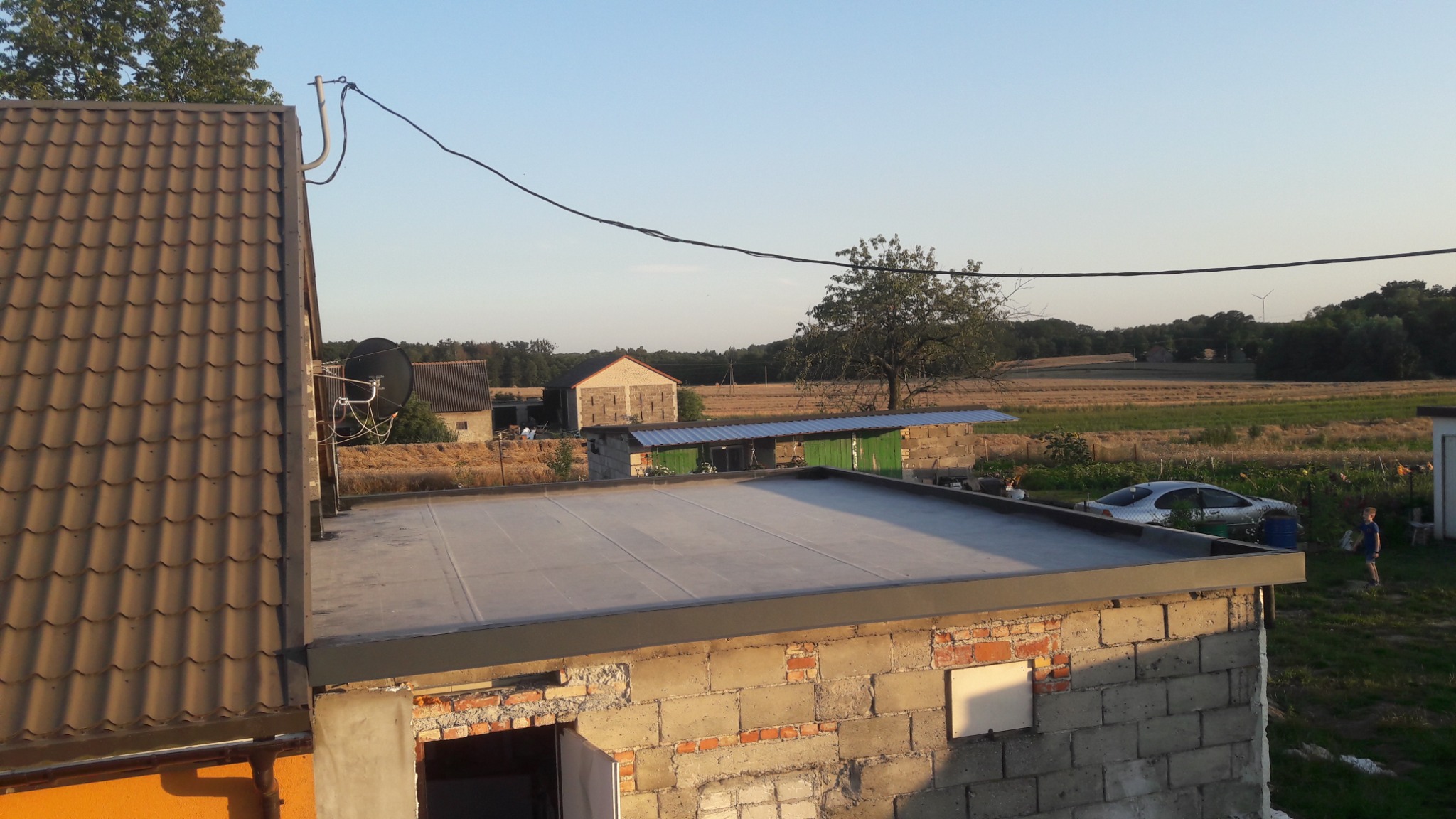 Widok z góry na płaski dach z papy na budynku w trakcie budowy, z widocznymi cegłami i otoczeniem wiejskim w tle, w tym pole, budynki gospodarcze i turbina wiatrowa w oddali.