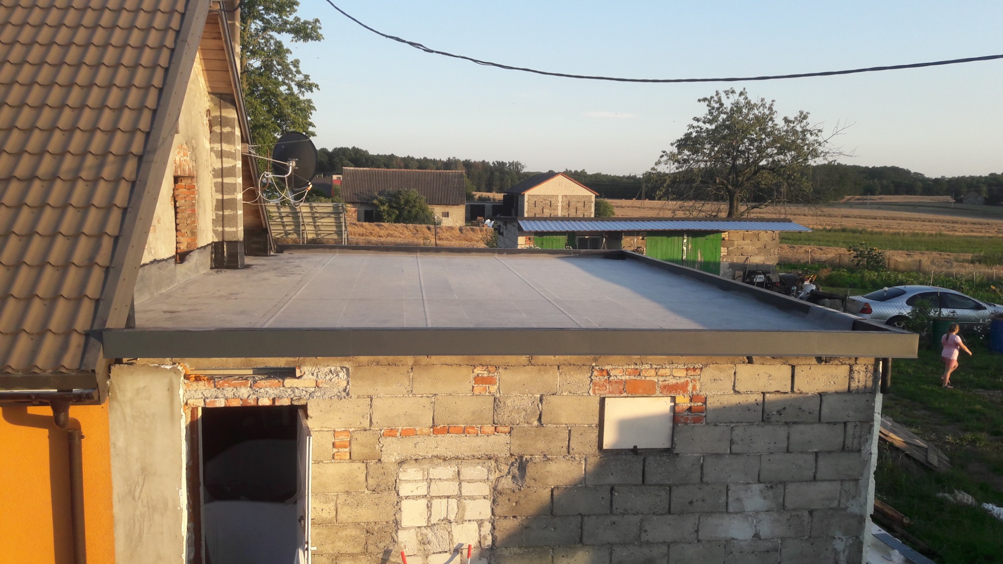 Wykończony płaski dach budynku gospodarczego z widocznymi detalami obróbki blacharskiej, na tle wiejskiego krajobrazu i częściowo wyremontowanej ściany z cegły.