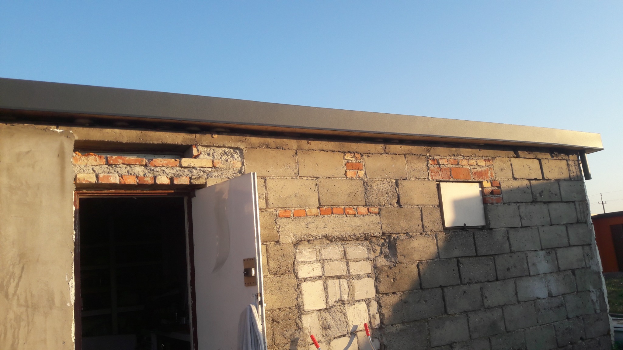 Surowy mur z betonowych bloków z fragmentami cegieł, częściowo otynkowany, z otwartymi drzwiami i nową metalową obróbką blacharską dachu w kolorze grafitowym.