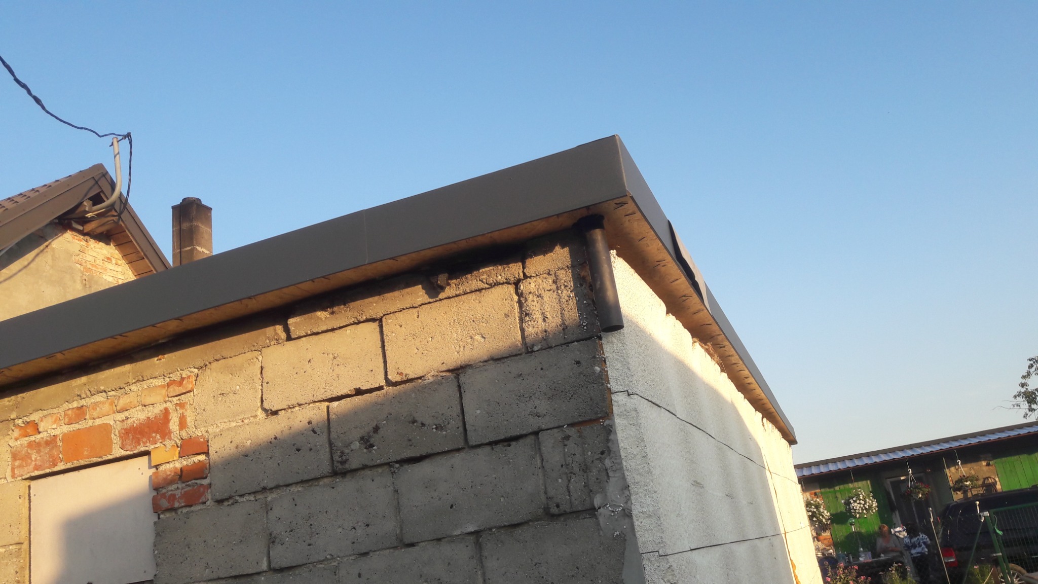 Narożnik budynku z szarej cegły z nową, grafitową obróbką blacharską, widoczne pozostałości starego dachu i komin w tle, oraz fragment sąsiedniego budynku.