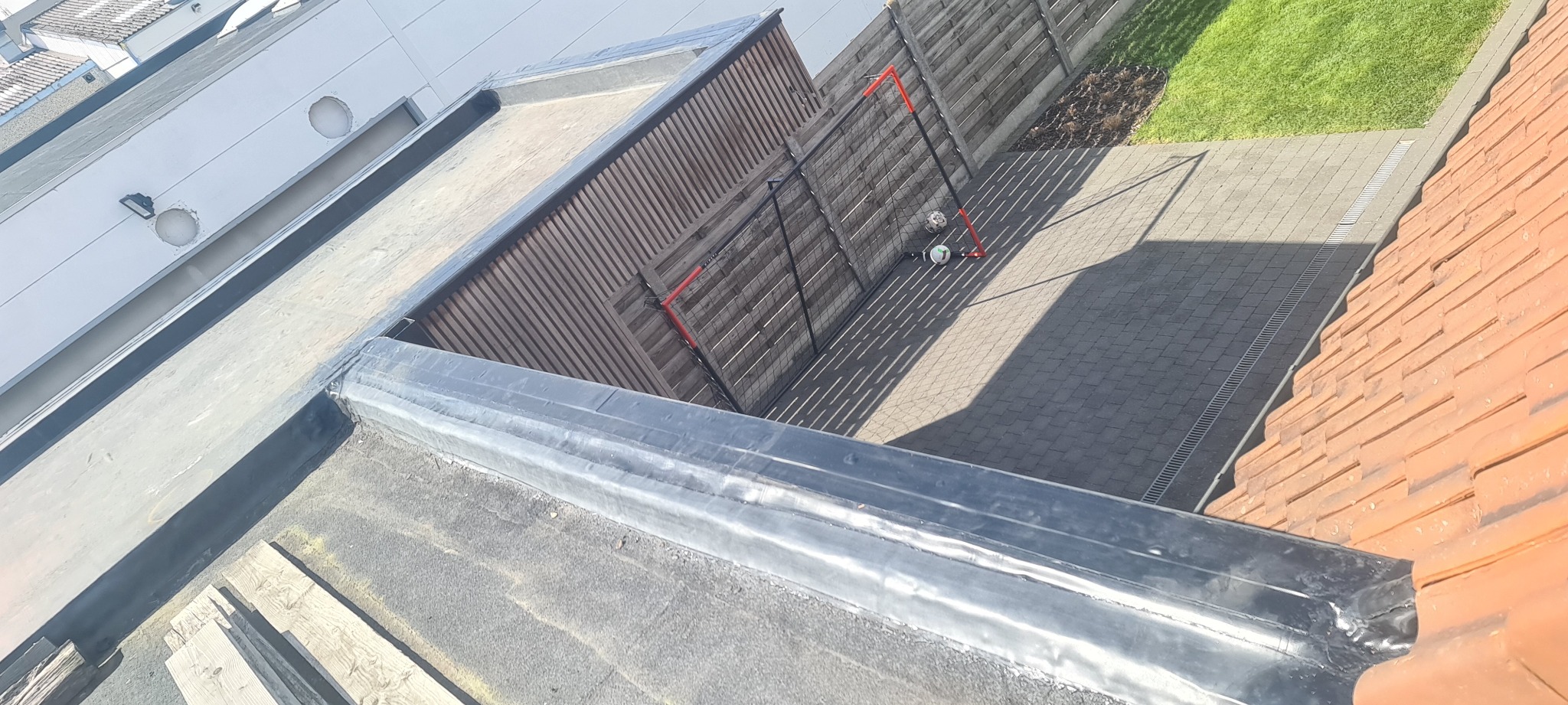 Widok z góry na płaski dach z obróbką blacharską, w tle ogrodzenie i fragment podwórka z bramką do piłki nożnej.
