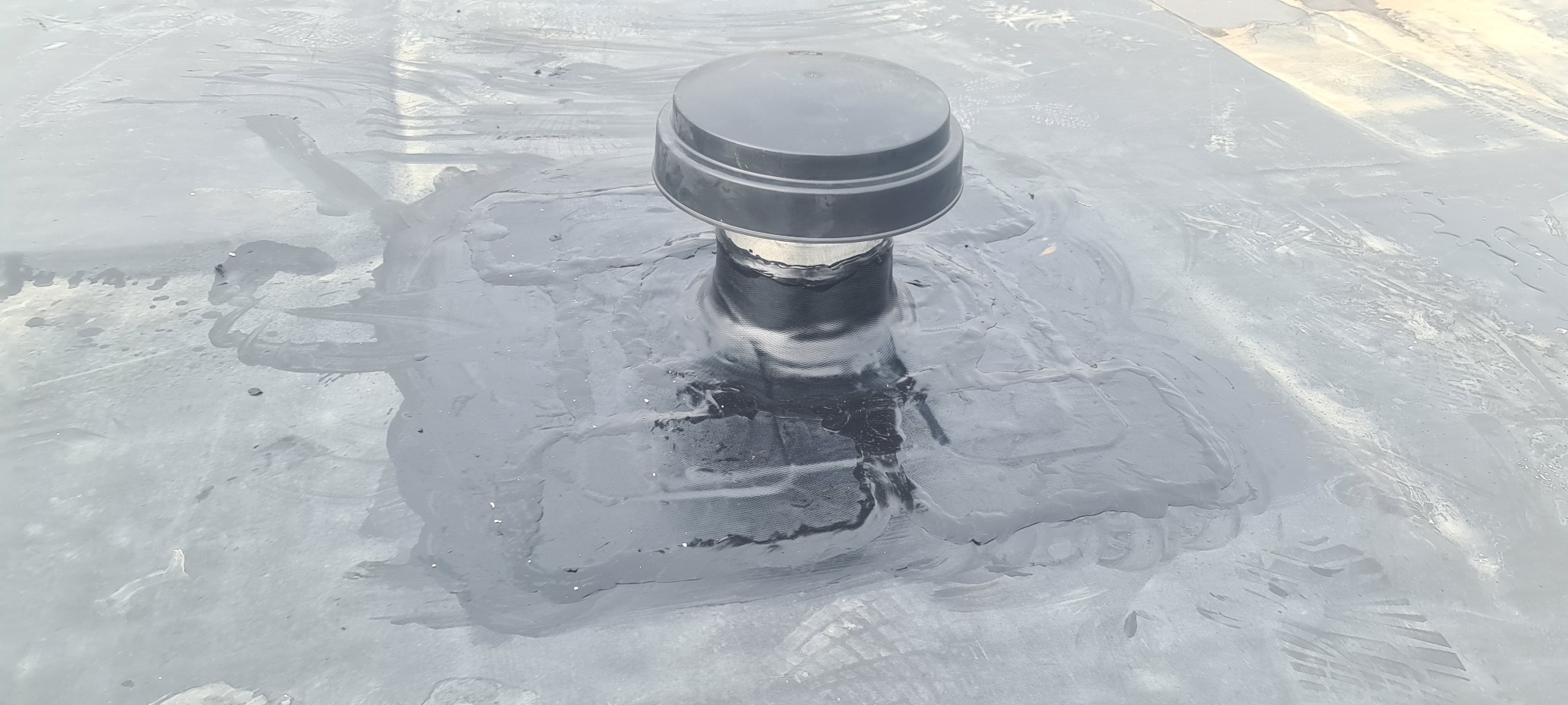 Czarny kominek wentylacyjny na płaskim dachu pokrytym papą, z widocznymi śladami świeżo nałożonej izolacji bitumicznej wokół podstawy.