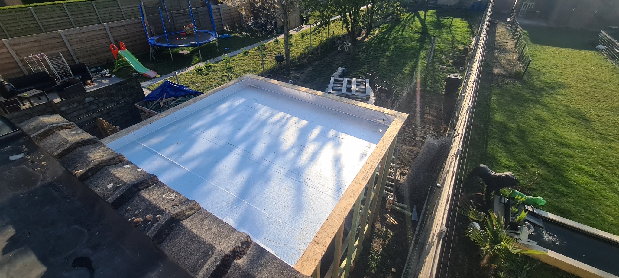 Płaski dach pokryty białą membraną, z drewnianą konstrukcją nośną, widok z góry na tle zielonego ogrodu z trampoliną i zjeżdżalnią dla dzieci.