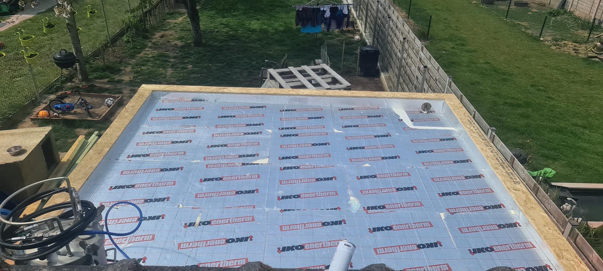 Płyty izolacyjne z folią aluminiową, marki IKO enertherm, na drewnianej konstrukcji dachu, widok z góry, z elementami narzędzi i ogrodu w tle.