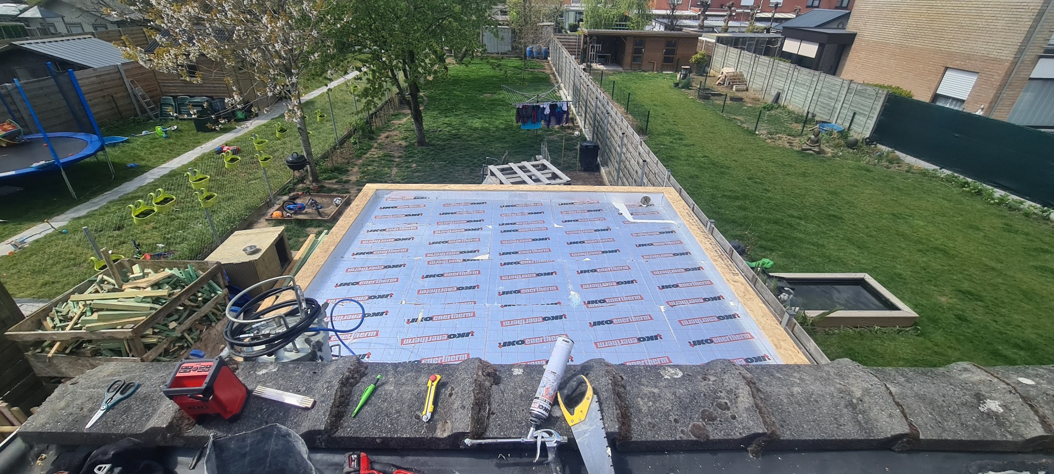 Widok z góry na przygotowywaną konstrukcję dachu z izolacją termiczną UKO Enertherm, narzędzia dekarza leżą na starych dachówkach, w tle ogród z trampoliną i suszącym się praniem.