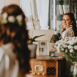 Arkadiusz Małecki Media - Panna młoda w szlafroku spogląda przez ramię, widoczna w lustrze, z bukietem białych kwiatów na pierwszym planie i regałem z książkami w tle.
