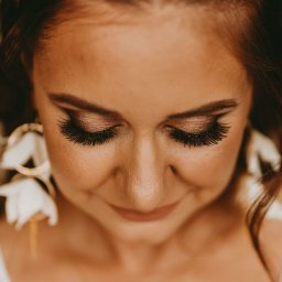 Arkadiusz Małecki Media - Portret kobiety z makijażem oczu w odcieniach brązu, długimi rzęsami i kolczykami w kształcie kwiatów, spoglądającej w dół.