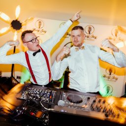 Arkadiusz Małecki Media - Dwóch mężczyzn w białych koszulach i muszkach pozuje za konsoletą DJ-ską podczas imprezy, jeden w okularach i szelkach, obaj z uniesionymi rękami, w tle ścianka z logo i dekoracyjne lampy...