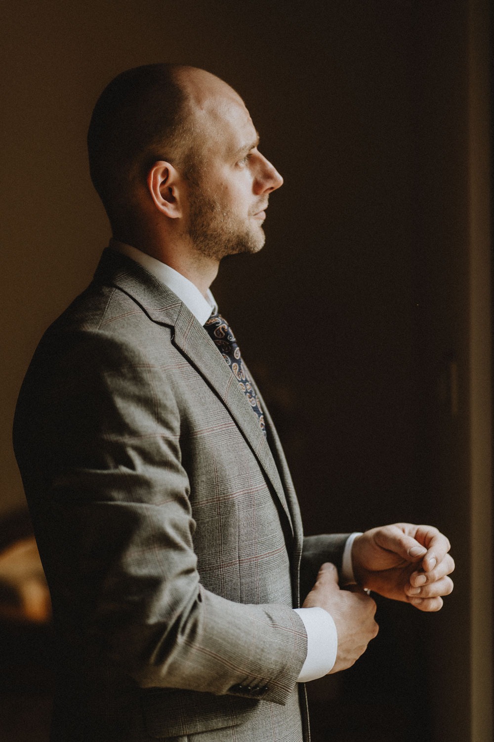 Mężczyzna w szarym garniturze w kratę, z wzorzystym krawatem, zapina mankiet koszuli przy oknie, profil.