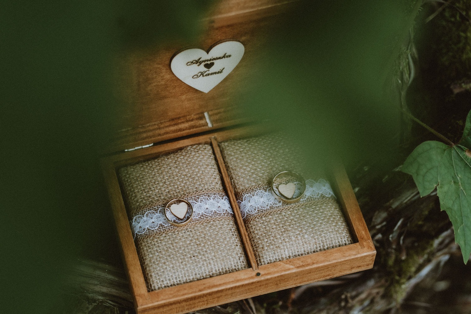 Otwarta drewniana szkatułka na obrączki z imionami Agnieszka i Kamil na serduszku, wypełniona jutą i koronką, z dwoma złotymi obrączkami z drewnianymi serduszkami w środku, widok z góry przez liście.