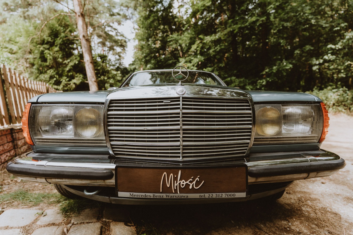 Przód klasycznego, szaro-niebieskiego Mercedesa z chromowanym grillem i napisem 'Miłość' na drewnianej tabliczce, zaparkowanego na tle zieleni.