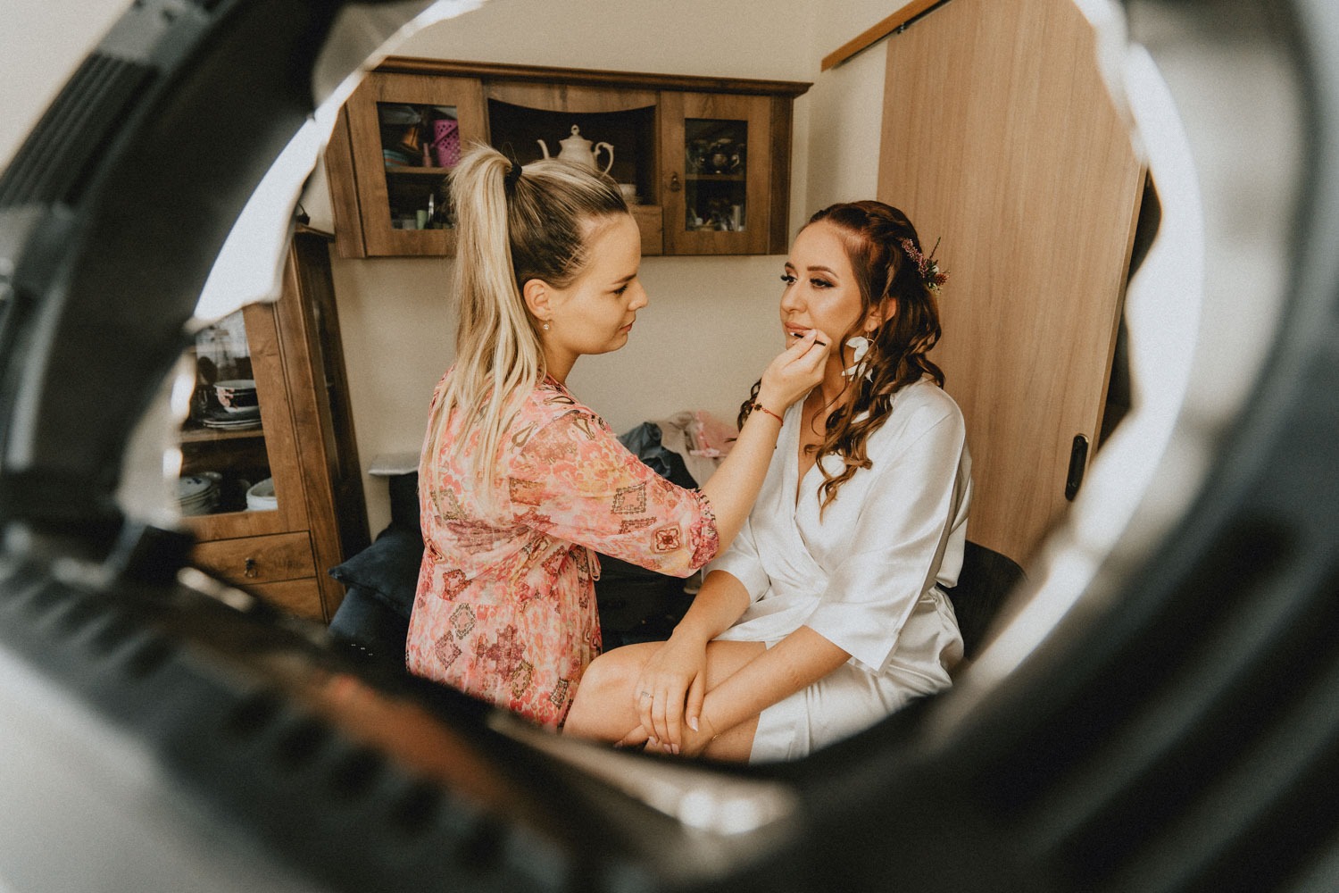 Kobieta w kwiecistej sukience nakłada szminkę pędzlem na usta siedzącej kobiety w białym szlafroku, widok przez pierścień lampy, w tle szafka i drzwi.