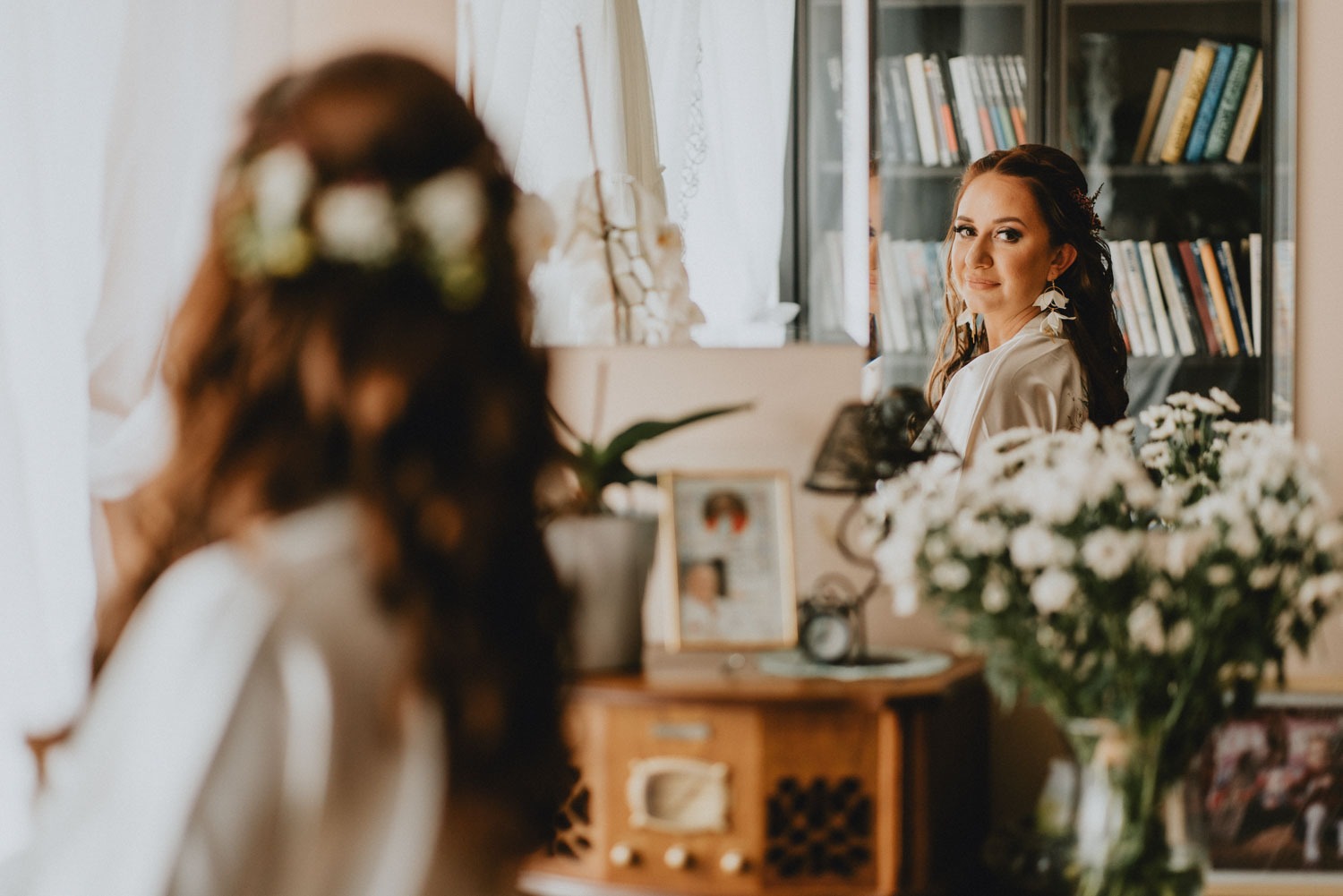 Panna młoda w szlafroku spogląda przez ramię, widoczna w lustrze, z bukietem białych kwiatów na pierwszym planie i regałem z książkami w tle.