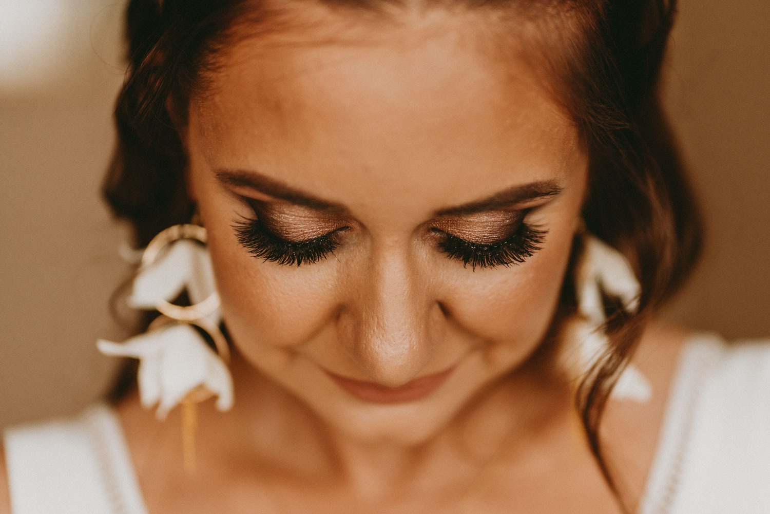 Portret kobiety z makijażem oczu w odcieniach brązu, długimi rzęsami i kolczykami w kształcie kwiatów, spoglądającej w dół.