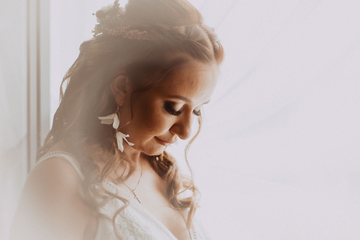 Portret kobiety z upiętymi włosami ozdobionymi kwiatami, w białej sukience z koronkowym wykończeniem, noszącej kolczyki w kształcie białych kwiatów i złoty łańcuszek z krzyżykiem, spoglądającej w dół.