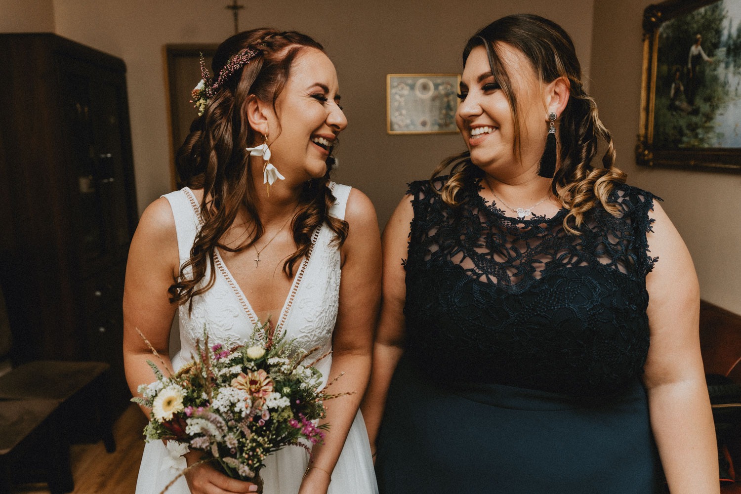 Uśmiechnięta panna młoda w białej sukni z bukietem polnych kwiatów patrzy na druhnę w granatowej koronkowej sukience.