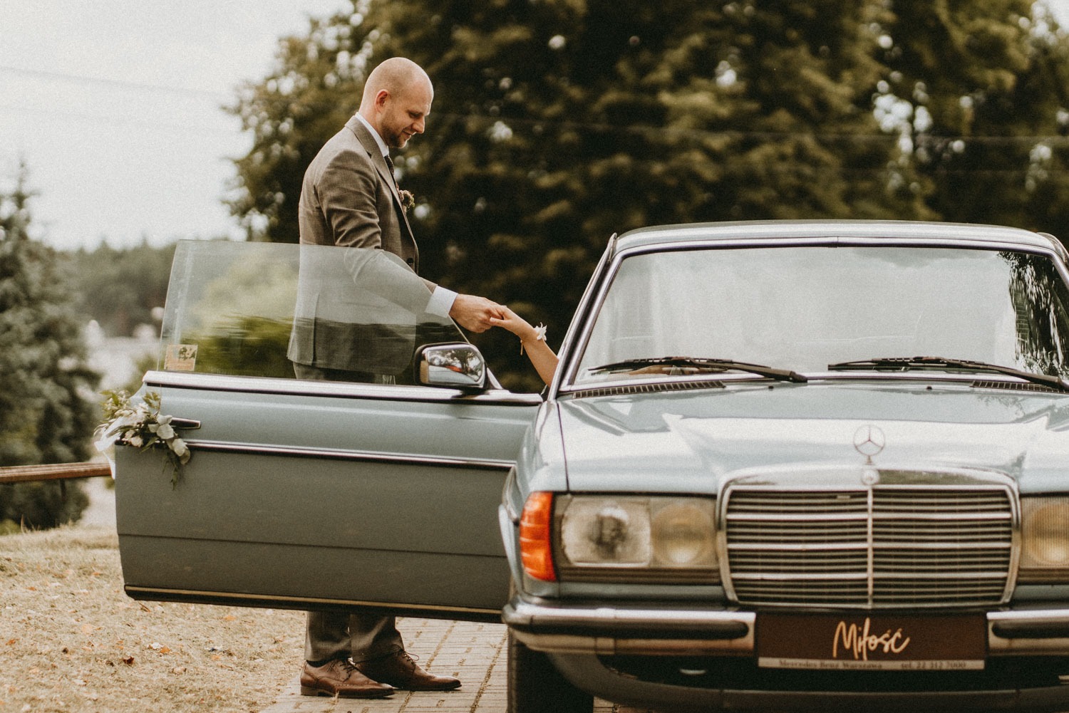 Panna młoda wychodząca z klasycznego, turkusowego Mercedesa, trzymana za rękę przez pana młodego w tweedowym garniturze, bukiet kwiatów na drzwiach auta.