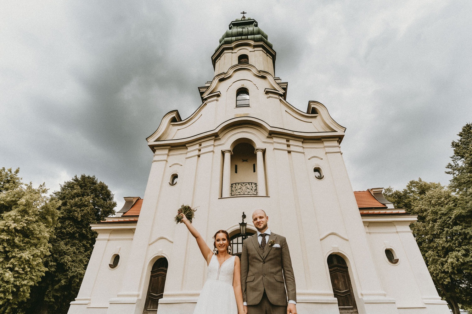Para młoda pozuje przed kościołem z wieżą zakończoną krzyżem, panna młoda trzyma bukiet kwiatów w uniesionej ręce, pochmurne niebo w tle.