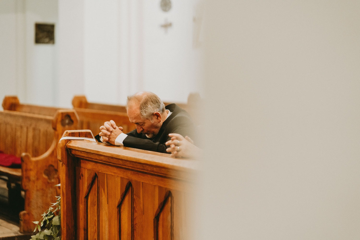 Starszy mężczyzna w ciemnym garniturze modli się w kościele, opierając czoło na drewnianej ławce, z rękami złożonymi w geście modlitwy, w tle widoczne inne ławki i fragment dekoracji.