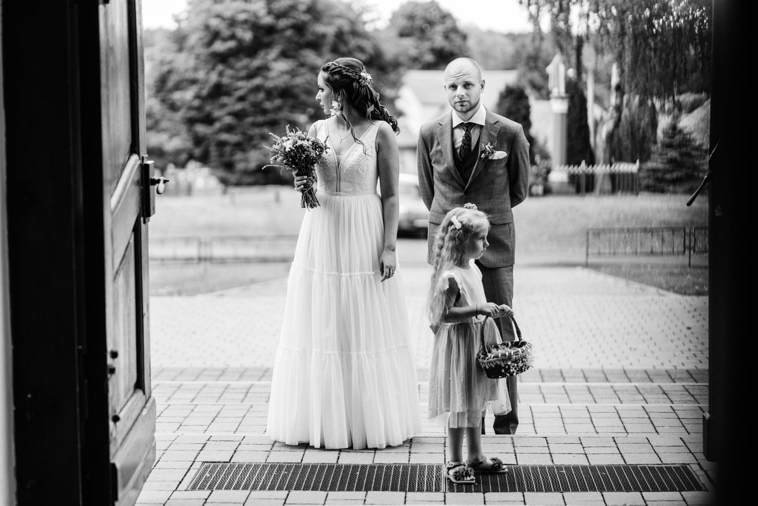 Czarno-białe zdjęcie panny młodej z bukietem, pana młodego i dziewczynki z koszyczkiem kwiatów stojących na schodach przed wejściem do budynku, z otwartymi drzwiami po lewej stronie.