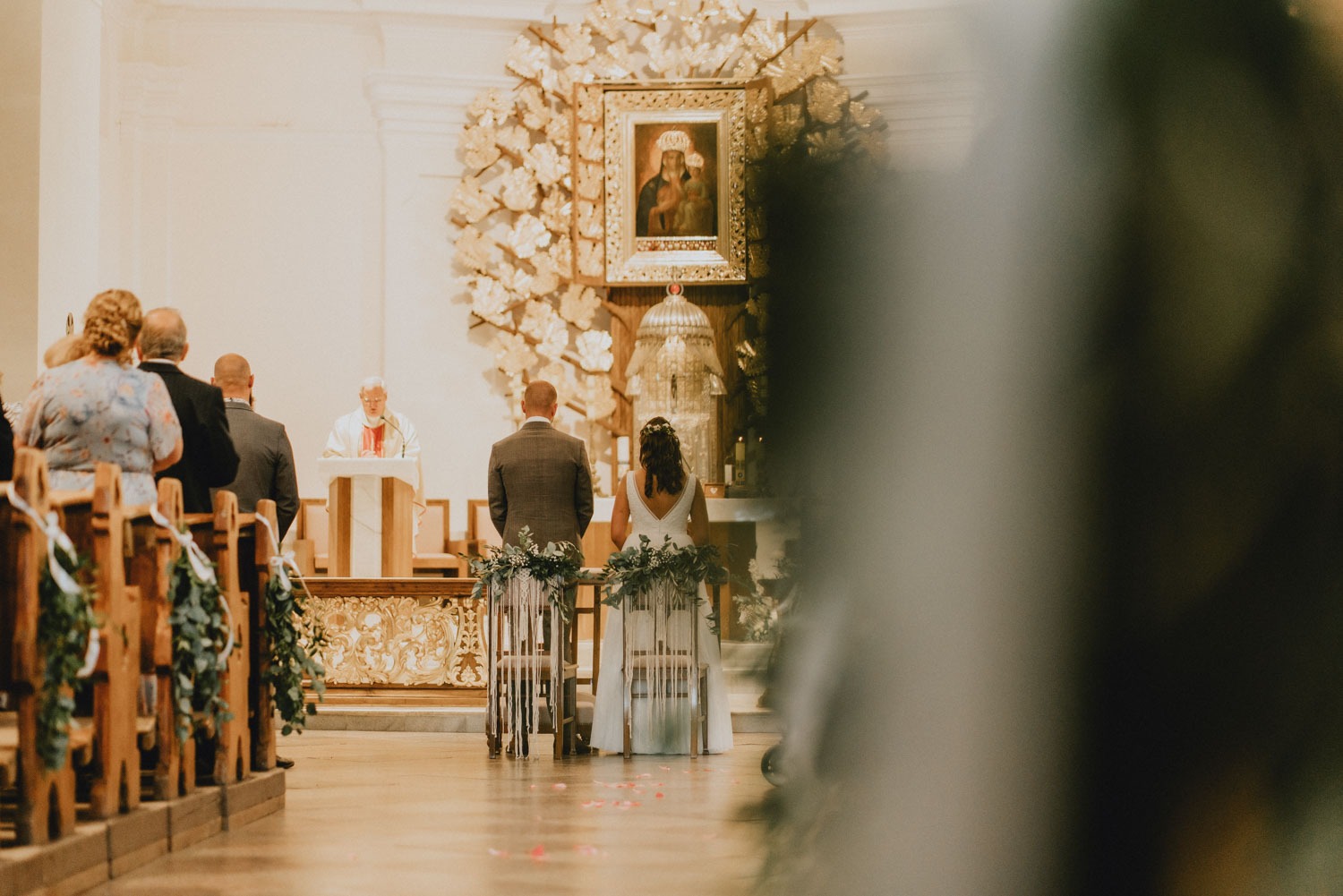 Para młoda stoi przed ołtarzem podczas ceremonii ślubnej w kościele, widok z tyłu, dekorowane krzesła, płatki róż na podłodze, ksiądz w tle.