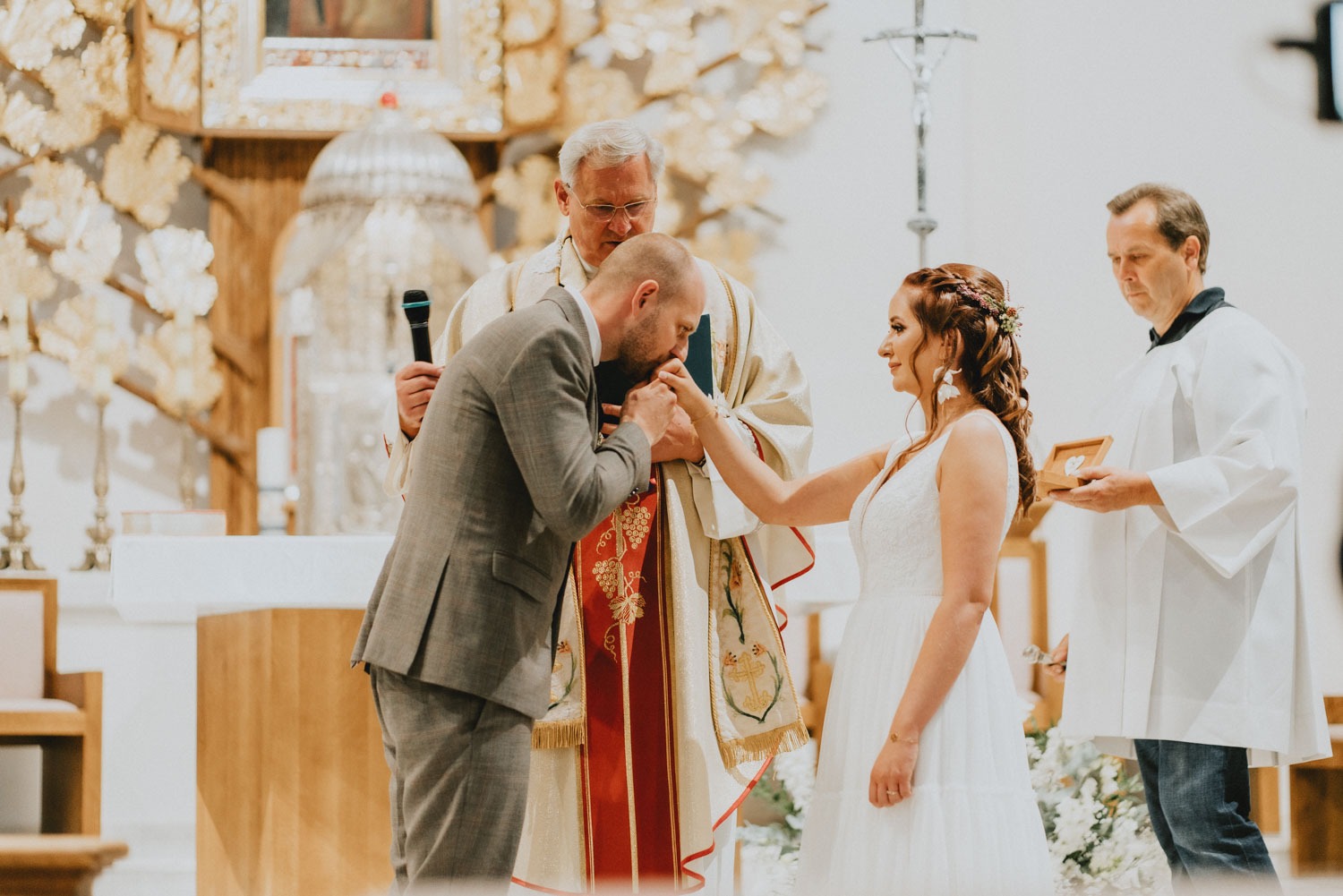 Pan młody w szarym garniturze całuje dłoń panny młodej w białej sukni ślubnej podczas ceremonii w kościele, w tle ksiądz w szatach liturgicznych i ministrant z poduszką na obrączki.