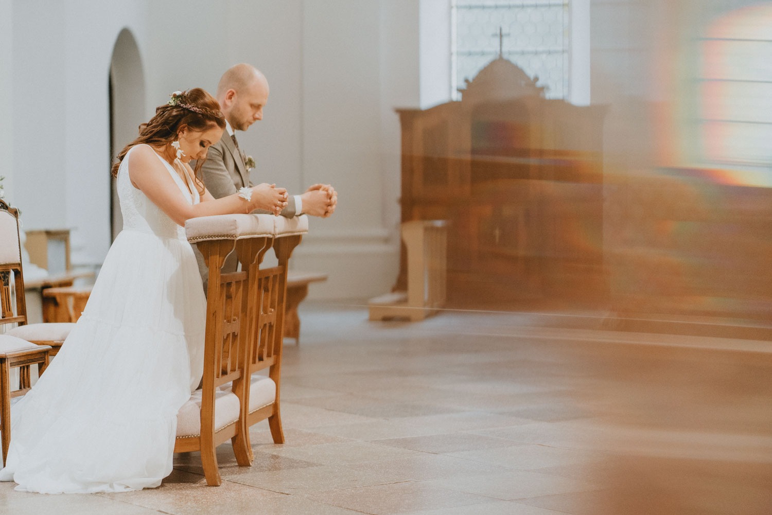 Para młoda w kościele, klęcząca w ławkach, z delikatnym światłem wpadającym przez okno, tworzącym tęczowe refleksy na ścianie.