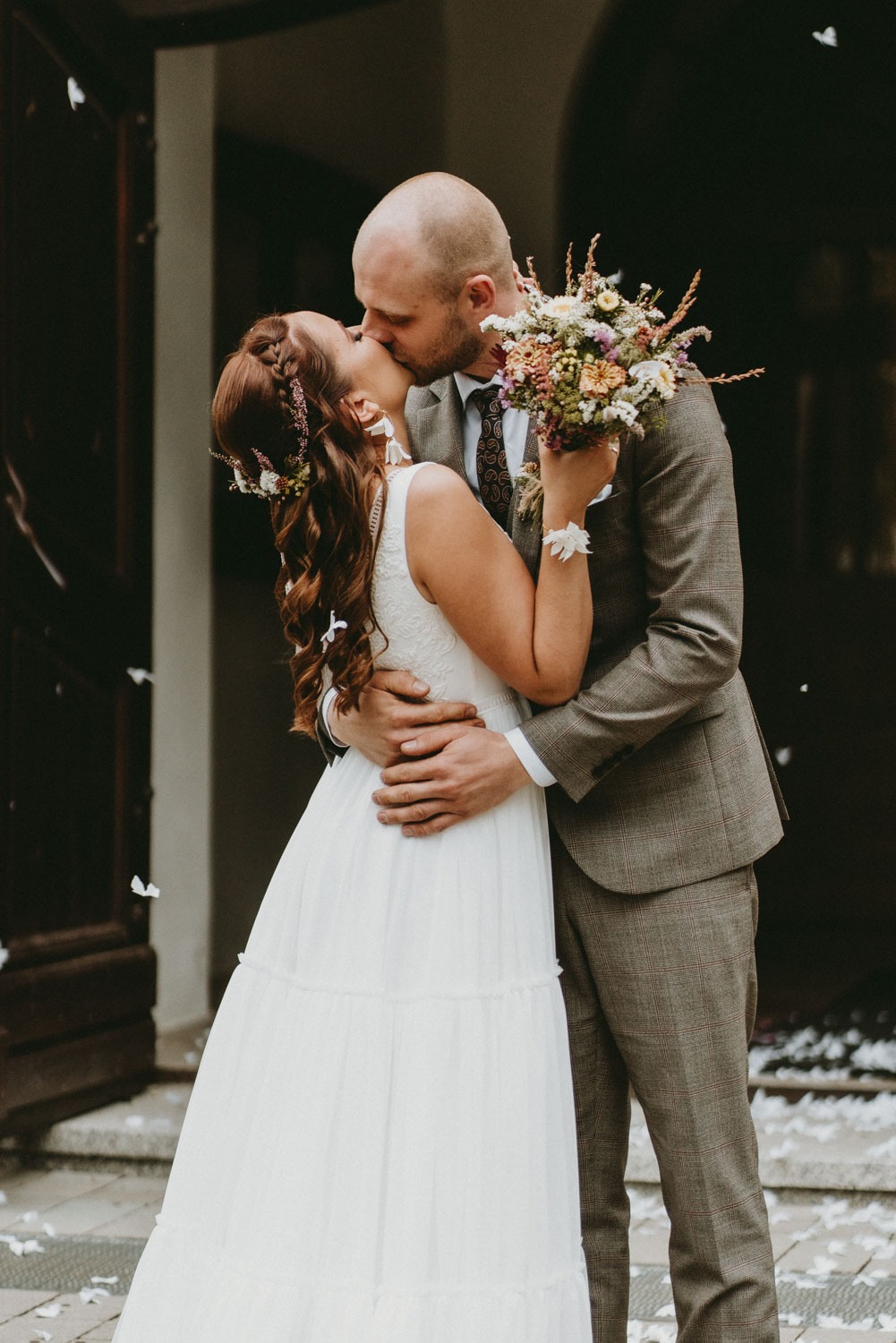 Pocałunek młodej pary przed wejściem do budynku, panna młoda trzyma bukiet polnych kwiatów, w tle opadające płatki kwiatów.