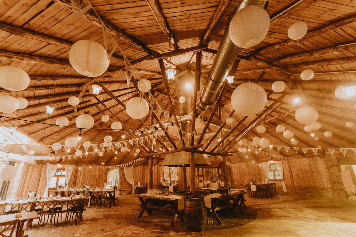 Drewniana sala z dekoracjami: papierowe lampiony, girlandy, drewniane stoły z kwiatami i krzesłami, ceglana podłoga, rustykalny klimat.