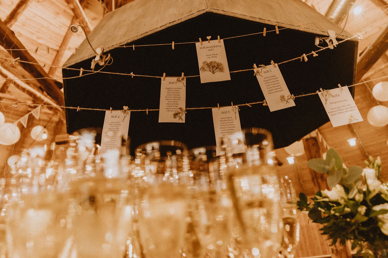 Dekoracja sali weselnej w Komornikach: tablica z listą gości przyczepiona klamerkami do sznurka, w tle sufit z drewnianych belek i wiszące lampiony, w pierwszym planie kieliszki i bukiet kwiatów.
