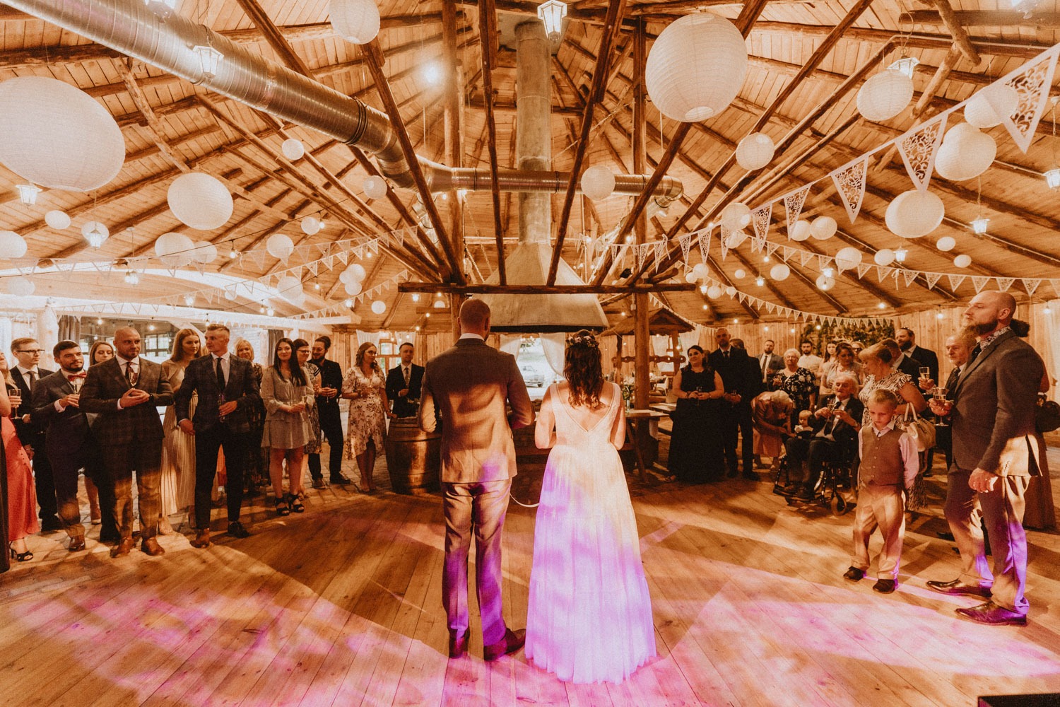 Para młoda stoi tyłem do widza na drewnianym parkiecie w rustykalnej stodole, otoczona gośćmi weselnymi, dekoracje z papierowych lampionów i girland.