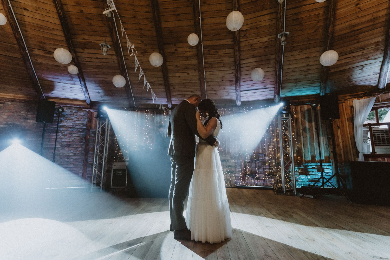 Para młoda podczas pierwszego tańca w rustykalnej sali weselnej z drewnianym stropem, oświetlonej reflektorami i girlandami świetlnymi.
