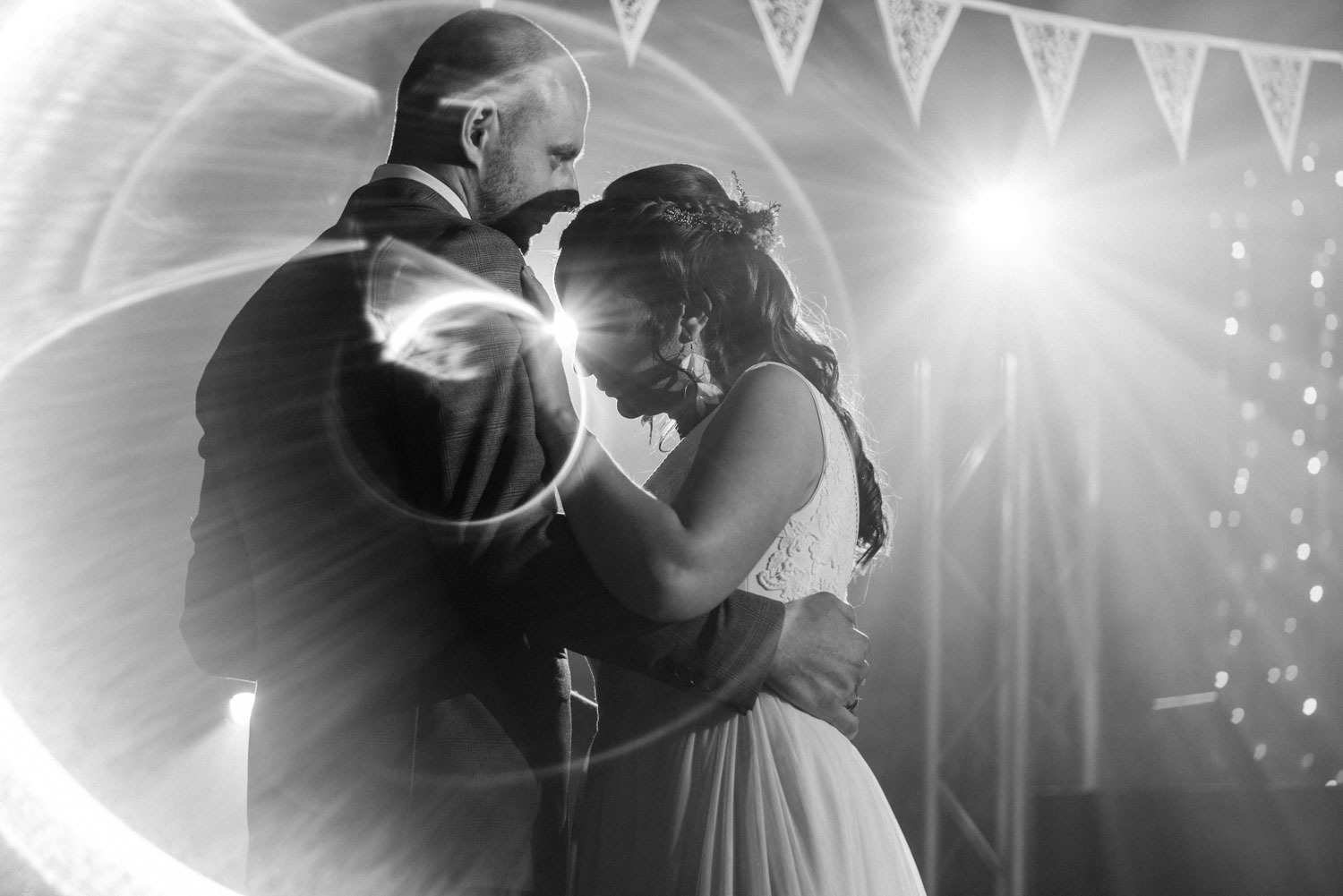 Czarno-białe zdjęcie pary młodej podczas pierwszego tańca, oświetlone mocnym światłem z tyłu, dekoracje w tle, efektowne rozmycie światła.