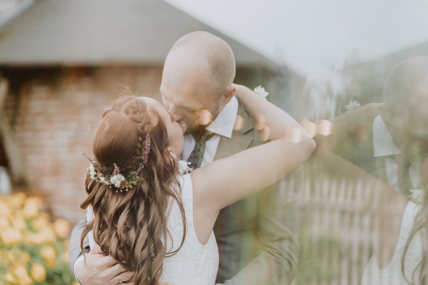 Pocałunek pary młodej w plenerze, panna młoda z wiankiem z kwiatów we włosach, pan młody w tweedowym garniturze z wzorzystym krawatem.