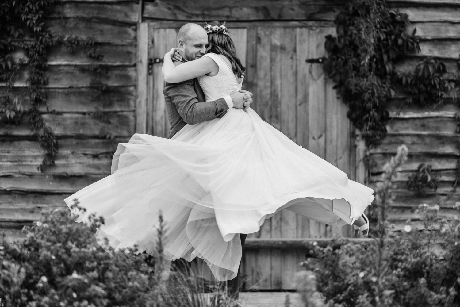 Czarno-białe zdjęcie wirującej panny młodej w sukni ślubnej, obejmowanej przez pana młodego na tle drewnianej stodoły porośniętej bluszczem.