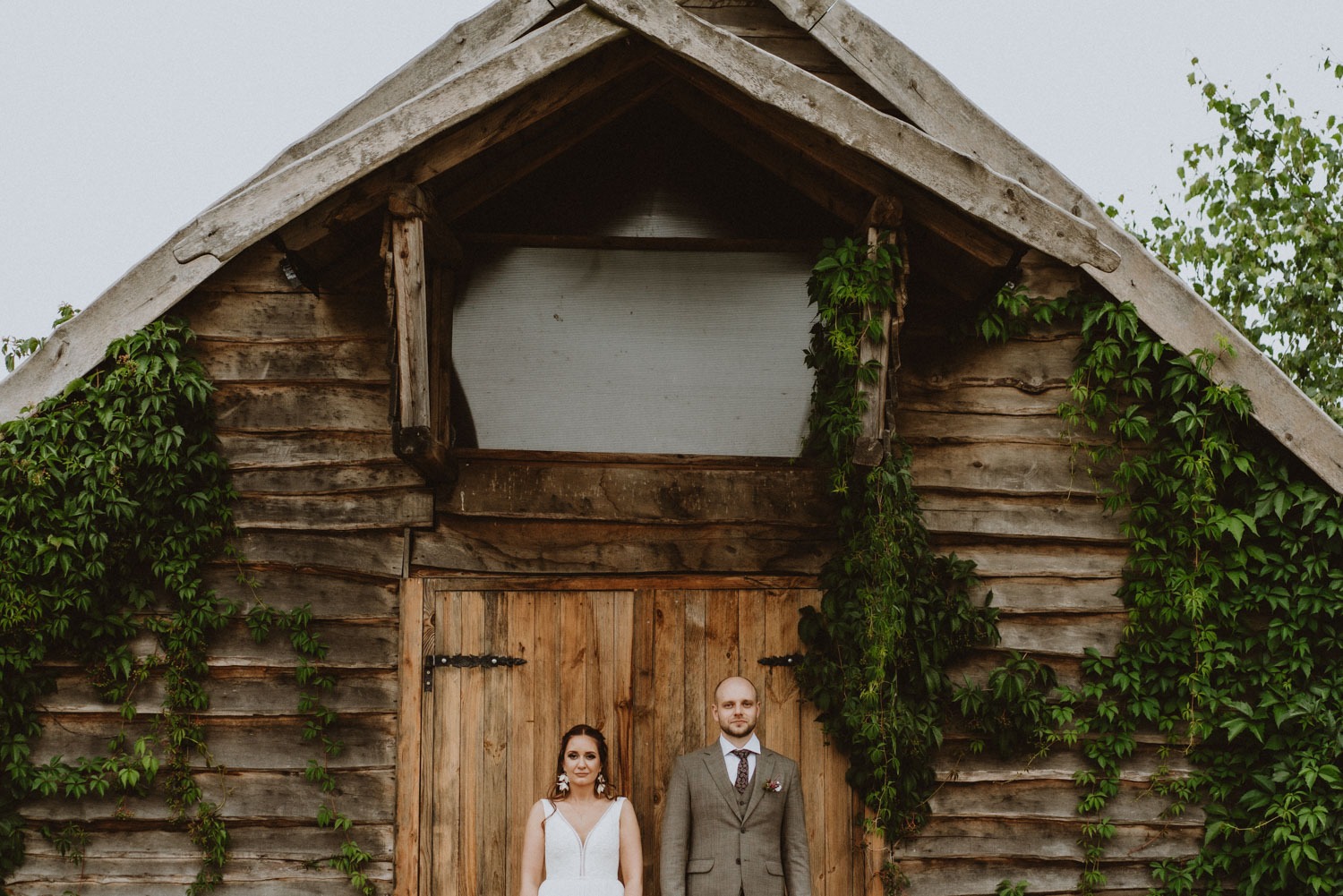 Para młoda stoi przed rustykalną, drewnianą stodołą z drzwiami i pnącą się zielenią, w tle szare niebo.