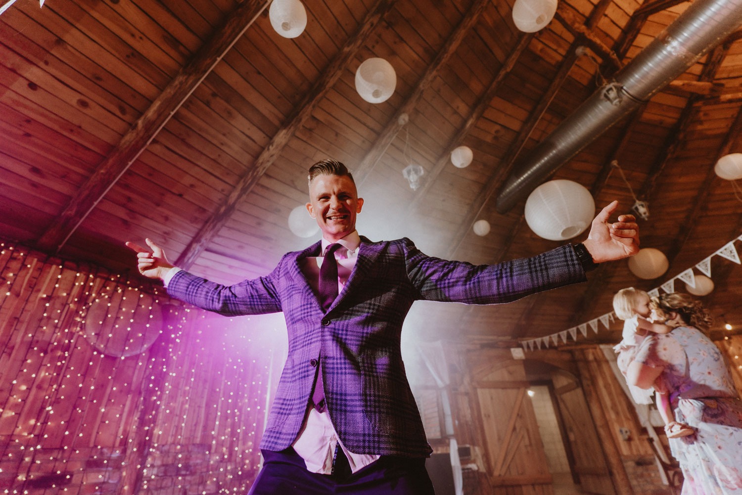 Uśmiechnięty mężczyzna w fioletowym garniturze w kratę z rozłożonymi ramionami na parkiecie sali weselnej z drewnianym stropem, lampionami i dekoracyjnym oświetleniem, w tle kobieta z dzieckiem.