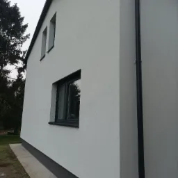 Wykończona elewacja domu jednorodzinnego w Żarkach, z widocznym fragmentem orynnowania i dwoma oknami z czarnymi ramami, wykończona na biało z ciemnym cokołem.