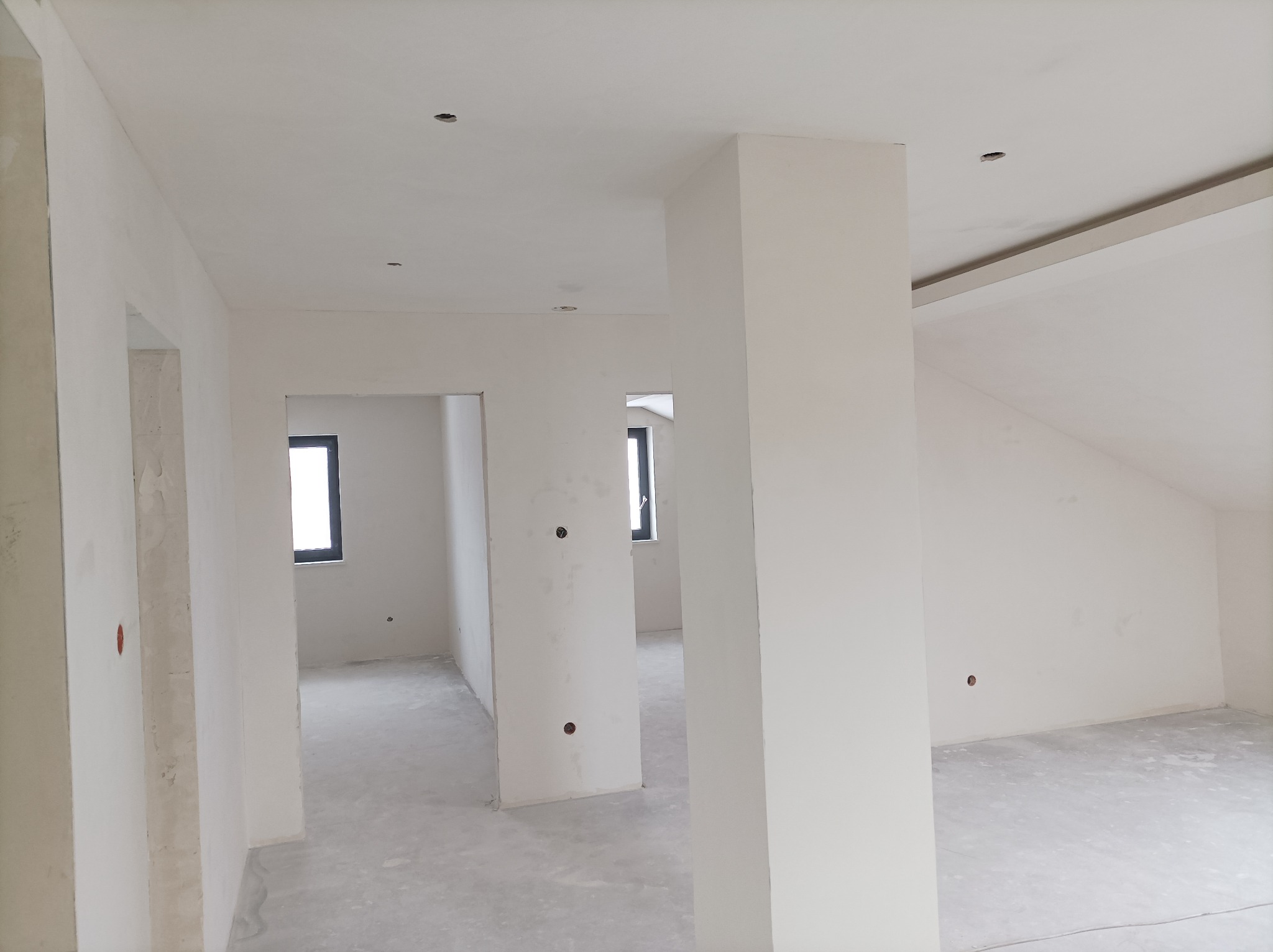 Wnętrze domu w stanie deweloperskim, widoczne białe ściany z otworami na gniazdka elektryczne, surowy beton na podłodze i okna z czarnymi ramami.