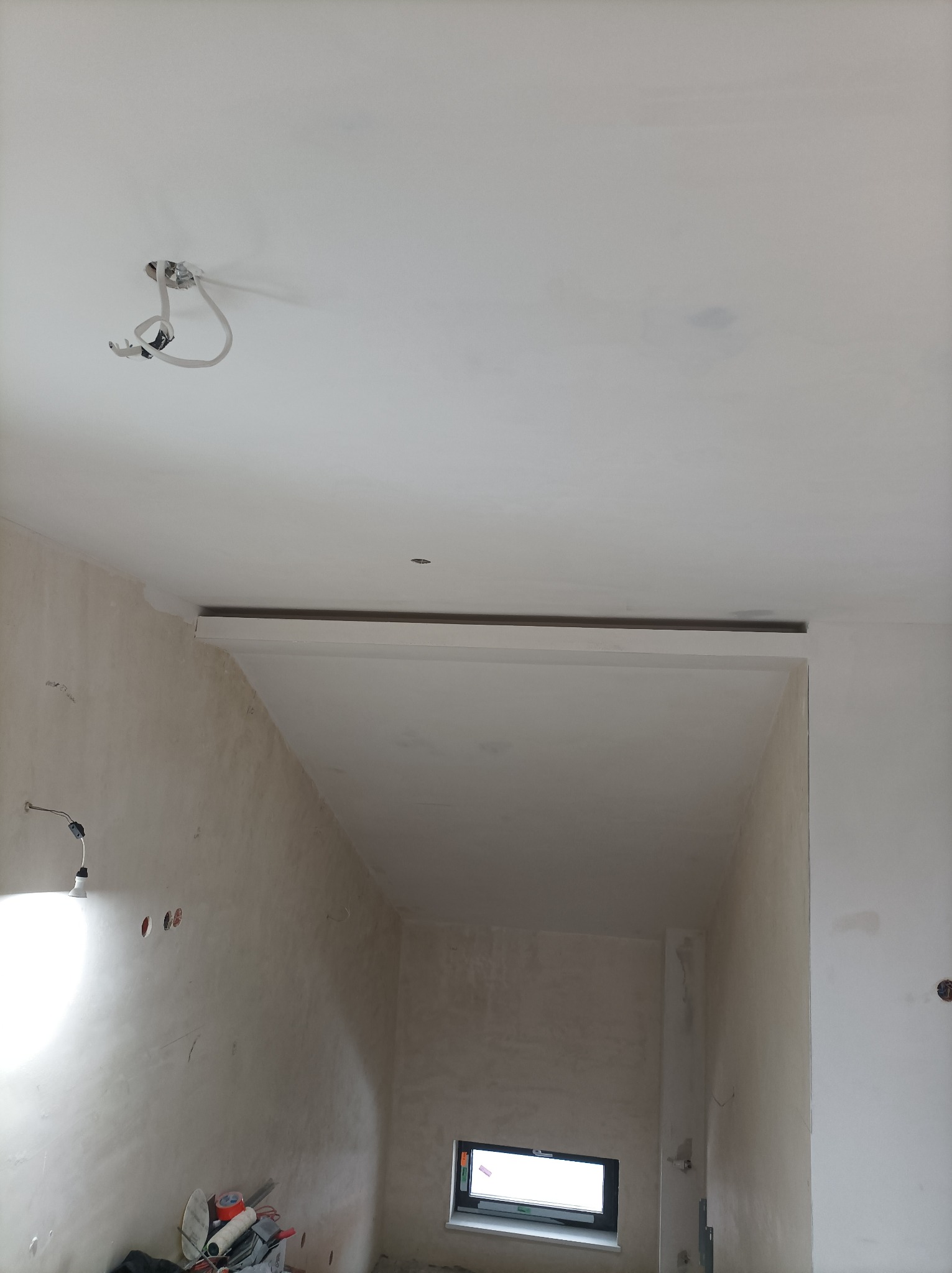 Wnętrze w trakcie prac wykończeniowych: surowe ściany, widoczne otwory na gniazdka, prowizoryczne oświetlenie z żarówką, okno w dolnej części kadru i narzędzia malarskie w rogu pomieszczenia.