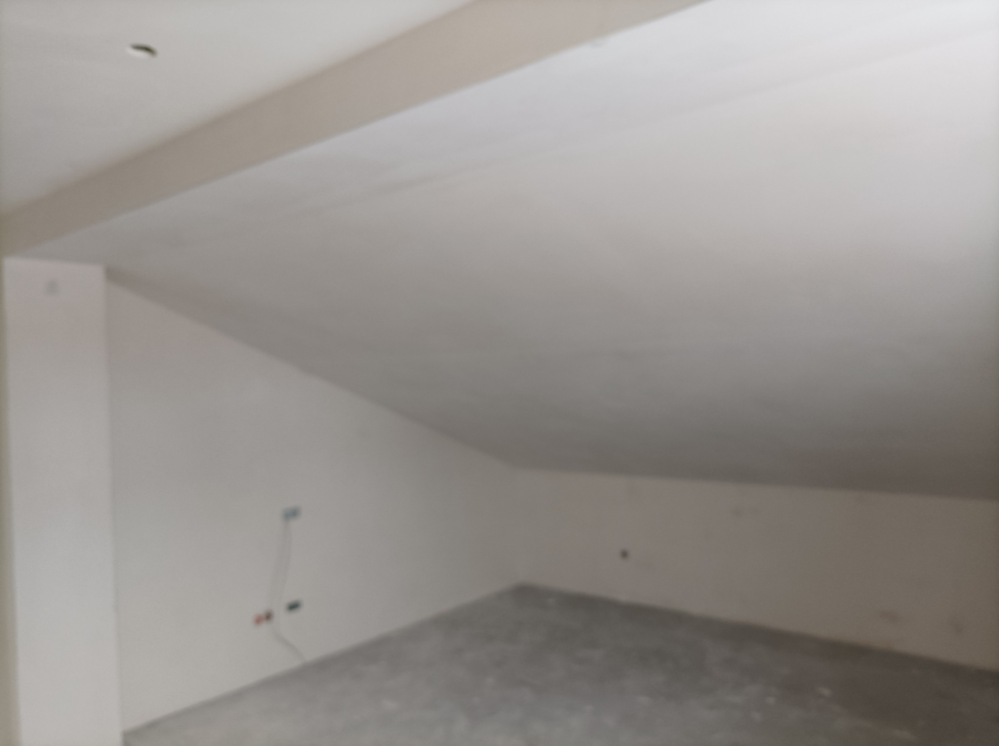 Wnętrze poddasza w trakcie prac wykończeniowych: otynkowane skośne ściany i sufit, widoczne wystające przewody elektryczne.