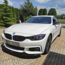 Białe BMW 4 Gran Coupe zaparkowane na kostce brukowej, z ciemnymi felgami, przyciemnionymi reflektorami, czarnym splitterem i elementami grilla w kolorach BMW Motorsport.