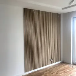 Ściana w pokoju z drewnianymi panelami ściennymi w pionowe paski, widoczne gniazdka elektryczne i drewniana podłoga parkietowa.