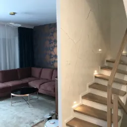 Salon z narożną kanapą, stolikiem kawowym, robotem sprzątającym i schodami z oświetleniem punktowym, ściana z efektem spękanego tynku.
