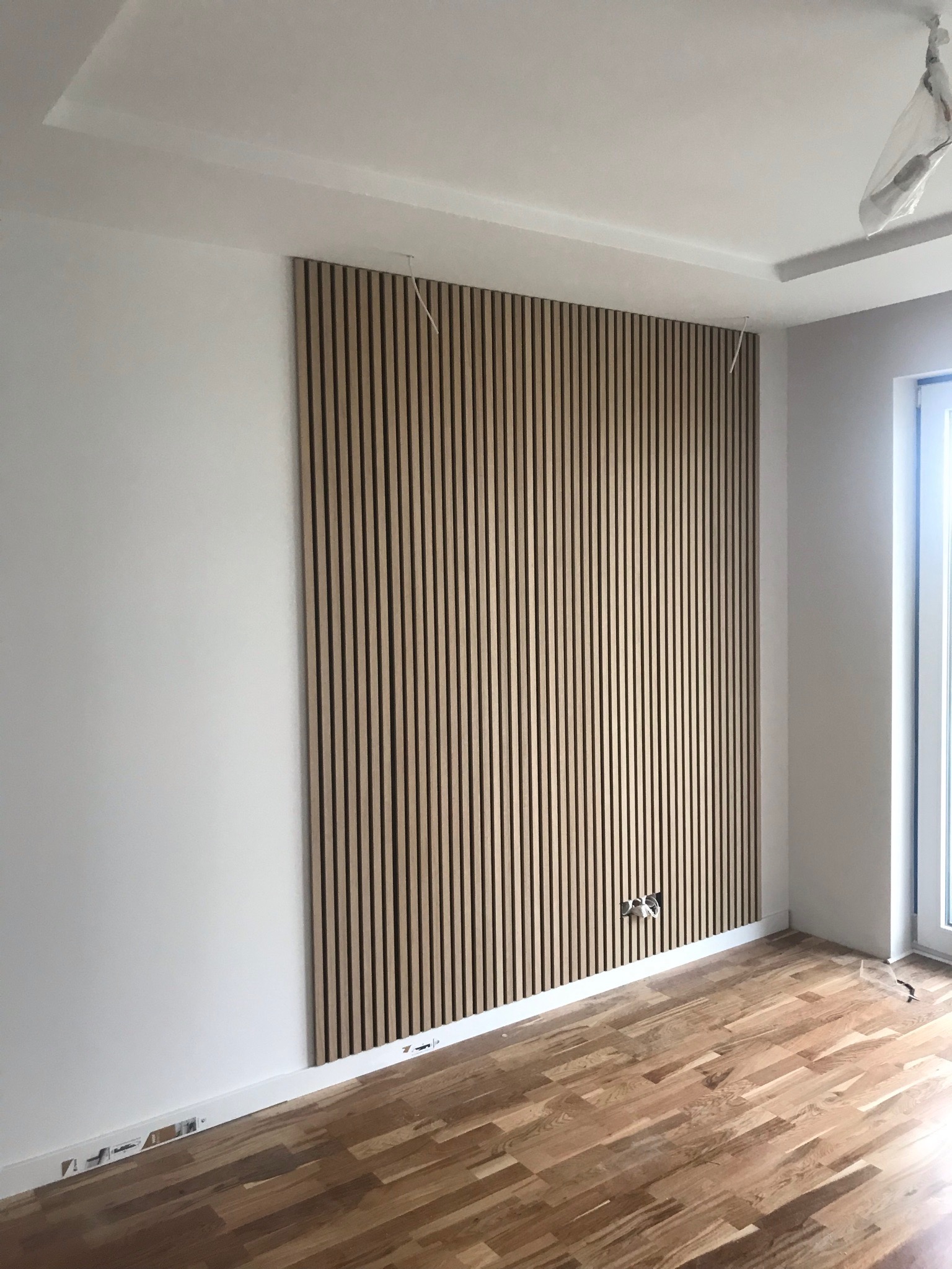 Ściana w pokoju z drewnianymi panelami ściennymi w pionowe paski, widoczne gniazdka elektryczne i drewniana podłoga parkietowa.