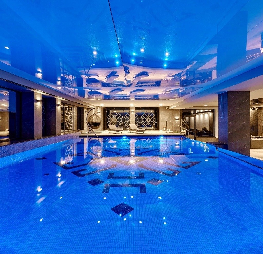 Kryty basen z mozaiką na dnie i sufitem z odbijającymi się elementami dekoracyjnymi, oświetlony niebieskim światłem, z leżakami i wiszącym fotelem w tle.